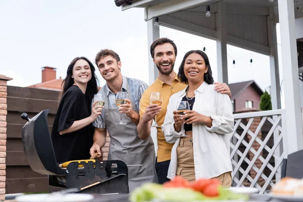 Sorrindo casais multiétnicos segurando copos de vinho e olhando para a câmera perto de comida na grelha no quintal — Fotografia de Stock