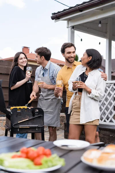 Homme positif étreignant ami bi-racial avec du vin tandis que les gens faisant barbecue — Photo de stock