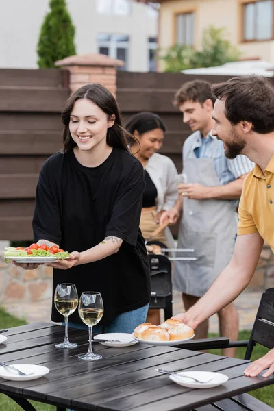 Donna positiva che tiene insalata vicino amico con panini durante il picnic in cortile — Foto stock