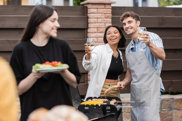 Lächelnde Freunde halten Wein in der Nähe von Essen auf dem Grill im Hinterhof — Stockfoto