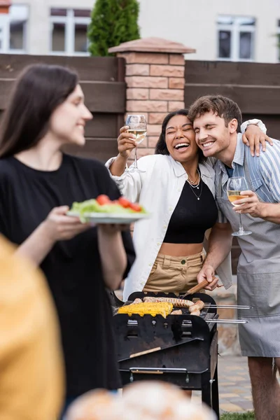 Feliz mulher bi-racial segurando vinho e abraçando amigo no avental fazendo churrasco ao ar livre — Fotografia de Stock