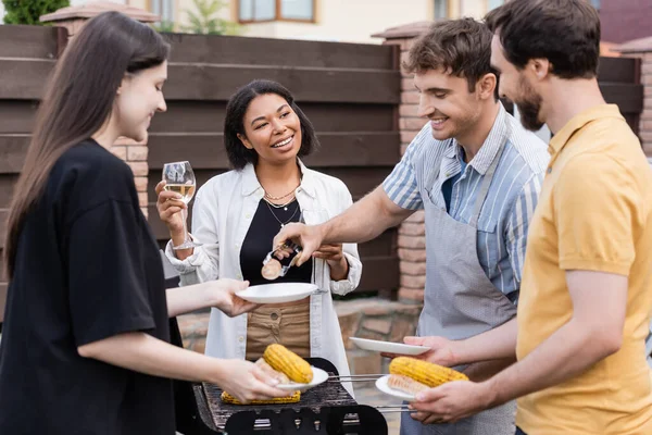 Man in apron putting grilled food on plates near multiethnic friends with wine — Stock Photo