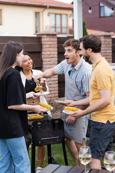 Shocked man in apron putting food on plates near interracial friends and blurred wine on backyard — Stock Photo