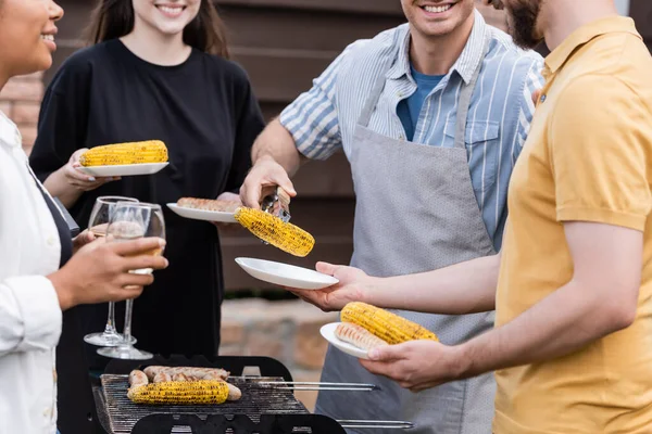 Обрізаний вид усміхненого чоловіка в фартусі, що тримає кукурудзу на грилі біля міжрасових друзів з тарілками та вином на відкритому повітрі — стокове фото