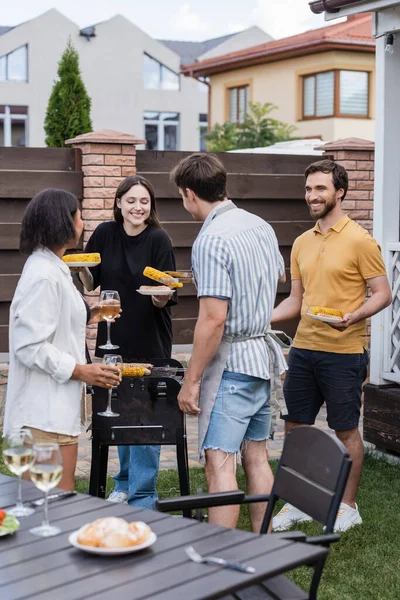 Amigos multiétnicos sonrientes con vino y comida en platos parados cerca de la parrilla en el patio trasero - foto de stock