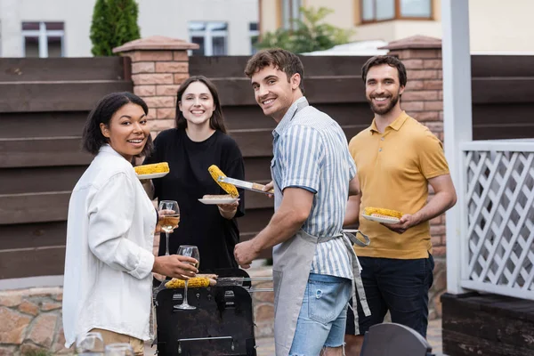 Amigos multiétnicos com vinho olhando para a câmera perto de comida grelhada ao ar livre — Fotografia de Stock