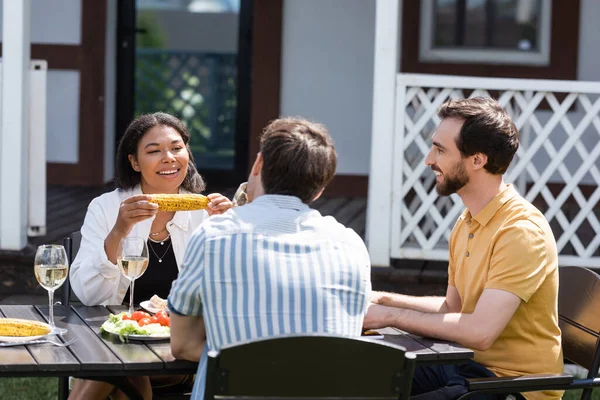 Cheerful multiethnic friends talking near grilled food and wine outdoors — Stock Photo