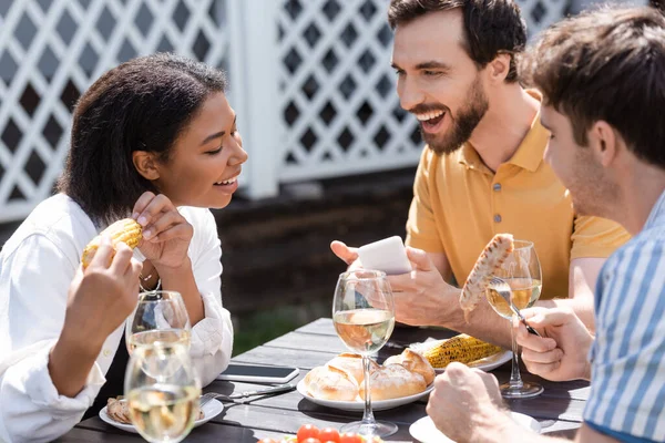 Homme souriant tenant smartphone et parlant à des amis interraciaux près de la nourriture et du vin pendant le pique-nique — Photo de stock