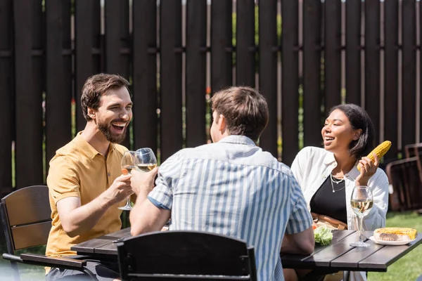 Cheerful men toasting with wine near bi-racial friend holding corn in backyard — Stock Photo