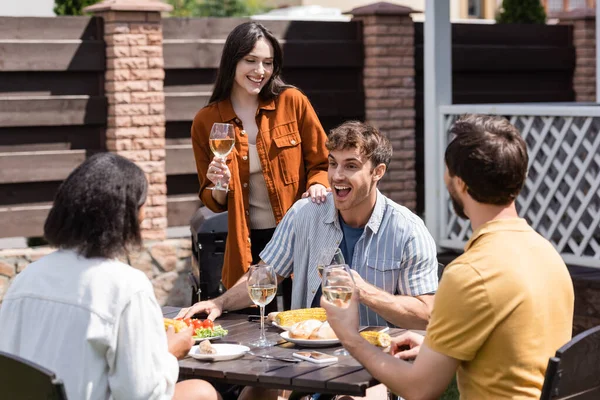 Positive interracial friends talking near tasty food and wine in backyard — Stock Photo