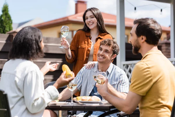 Позитивна жінка тримає келих вина поблизу розмитих друзів, які розмовляють під час пікніка на задньому дворі — стокове фото