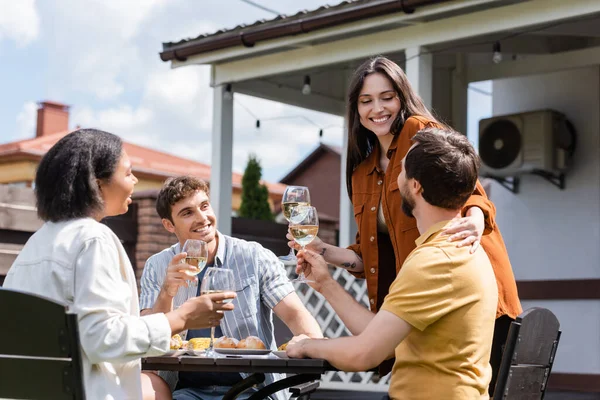Молодая женщина держит вино и обнимает мужчину рядом с межрасовыми друзьями во время пикника на заднем дворе — стоковое фото