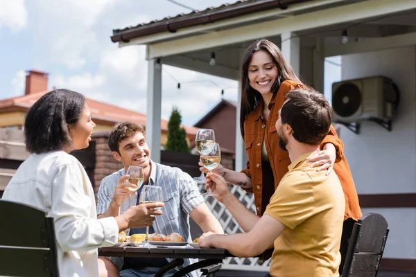 Mujer joven sosteniendo vino y abrazando al hombre cerca de amigos interracial durante el picnic en el patio trasero - foto de stock