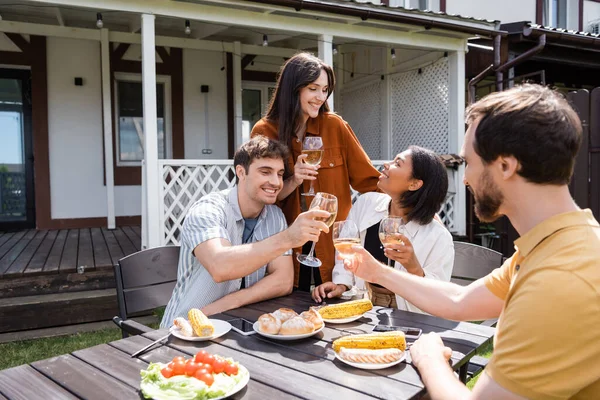 Smiling interracial friends holding wine near delicious grilled food in backyard — Stock Photo