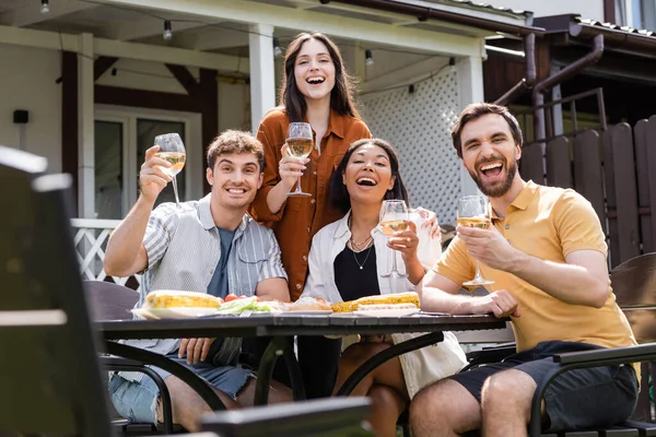 Amigos multiétnicos felizes com vinho olhando para a câmera perto de comida no quintal — Fotografia de Stock