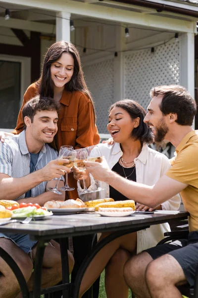 Allegri amici multietnici brindano con vino vicino al cibo alla griglia durante il picnic all'aperto — Foto stock