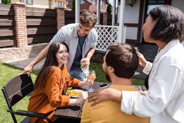 Positive woman holding glass of wine near boyfriend and interracial friends during picnic outdoors — Stock Photo