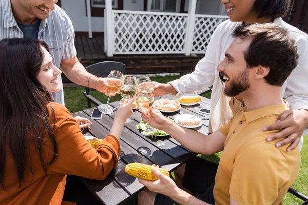 Amis interracial positifs griller avec du vin près de la nourriture dans la cour arrière de la maison — Photo de stock