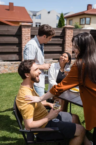 Femme souriante touchant l'homme avec du vin près des amis interracial flous pendant le pique-nique — Photo de stock