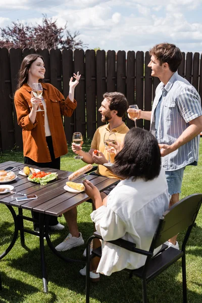 Mujer sonriente sosteniendo vino y hablando con amigos interracial durante el picnic en el patio trasero - foto de stock