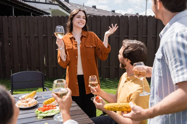 Веселая женщина, держащая вино и говорящая тосты за размытых многонациональных друзей во время пикника — стоковое фото