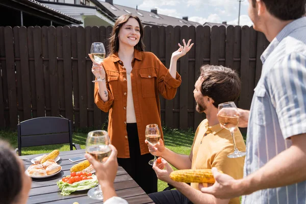 Femme joyeuse tenant du vin et parlant de pain grillé à des amis multiethniques flous pendant le pique-nique — Photo de stock