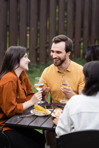 Homem positivo segurando copo de vinho perto de amigos inter-raciais e comida durante o piquenique — Fotografia de Stock