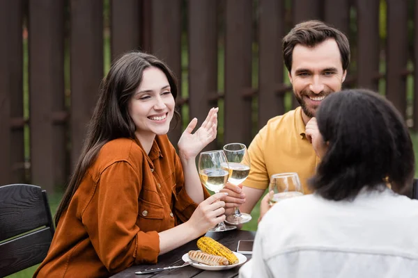Усміхнена жінка тримає вино і розмовляє з багатоетнічними друзями поблизу смаженої їжі та смартфона на відкритому повітрі — стокове фото