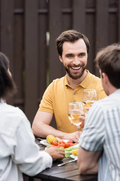 Smiling man holding glass of wine near blurred interracial friends and tasty grilled food outdoors — Stock Photo