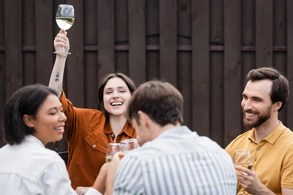 Fröhliche Frau hält ein Glas Wein in der Hand und blickt in die Kamera in der Nähe verschwommener interrassischer Freunde im Freien — Stockfoto