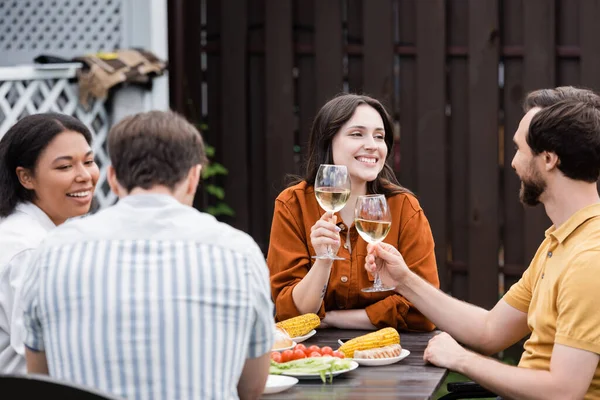 Gente positiva sosteniendo vino mientras borrosa amigos interracial hablando cerca de sabrosa comida al aire libre - foto de stock