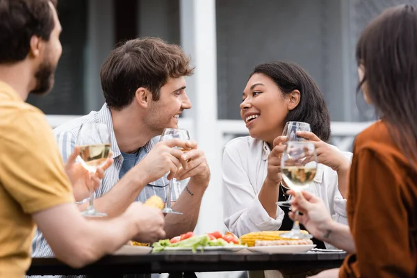 Glückliche bi-rassische Frau hält ein Glas Wein in der Hand und schaut tätowierte Freundin während einer Grillparty an — Stockfoto