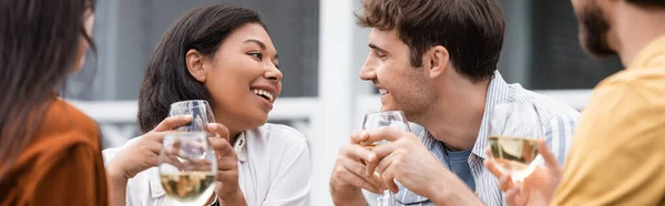 Feliz mulher bi-racial segurando copo de vinho e olhando para o amigo no quintal, banner — Fotografia de Stock