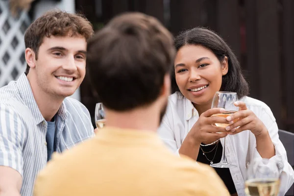 Happy multiethnic couple with glasses of wine looking at friend on blurred foreground — Stock Photo