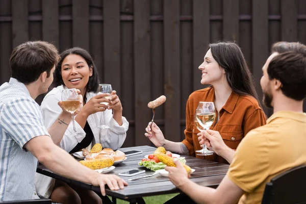 Fröhliche multiethnische Freunde halten Gläser mit Wein und essen während der Grillparty im Hinterhof — Stockfoto