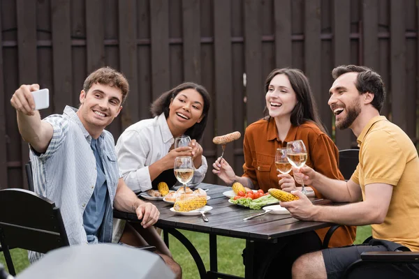 Alegres amigos multiétnicos tomando selfie durante la barbacoa en el patio trasero - foto de stock