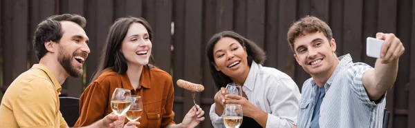 Alegres amigos multiétnicos tomando selfie durante la fiesta de barbacoa en el patio trasero, bandera - foto de stock