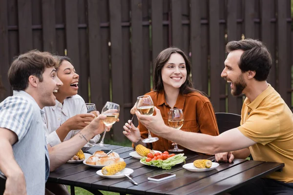 Cheerful multiethnic friends holding glasses with wine and having lunch during bbq party on backyard — Stock Photo