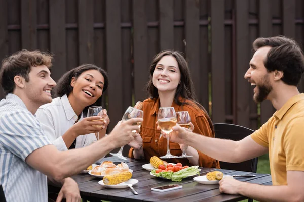 Alegres amigos multiétnicos tintinear vasos con vino y almorzar durante la fiesta de barbacoa - foto de stock