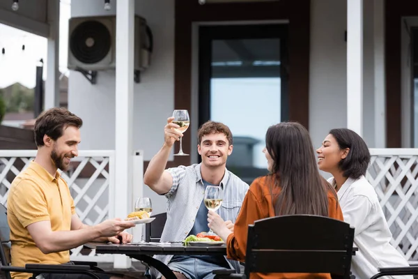 Happy man raising glass of wine near interracial friends during bbq party on backyard — Stock Photo