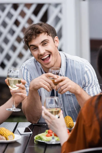 Hombre emocionado sosteniendo copa con vino cerca de amigos durante la fiesta de barbacoa en el patio trasero - foto de stock