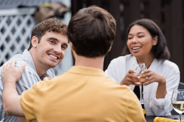 Cheerful multiethnic friends smiling near blurred man on foreground during bbq party on backyard — Stock Photo