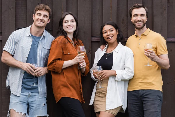 Happy interracial friends holding glasses with wine while standing near fence on backyard — Stock Photo