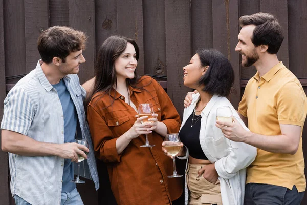 Happy interracial friends holding glasses with wine while smiling near fence on backyard — Stock Photo