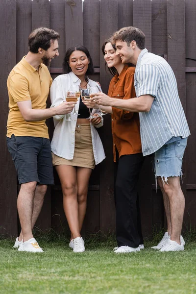 Full length of happy interracial friends clinking glasses with wine while standing near fence on backyard — Stock Photo