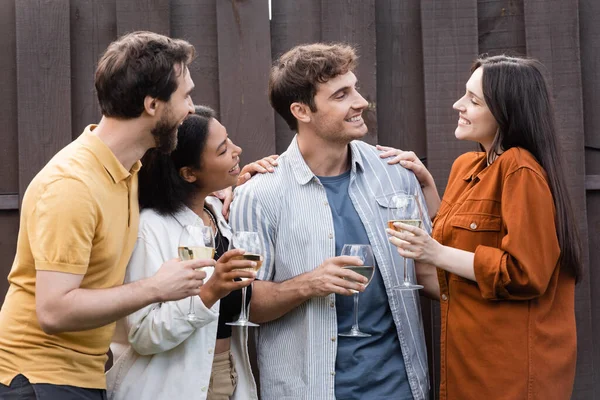 Cheerful interracial friends holding glasses with wine while smiling near fence on backyard — Stock Photo