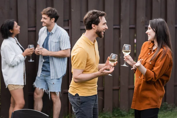 Cheerful couple holding glasses with wine while talking near blurred interracial friends on blurred background — Stock Photo
