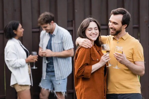 Casal alegre segurando copos com vinho enquanto abraçando perto de amigos inter-raciais borrados no fundo borrado — Fotografia de Stock