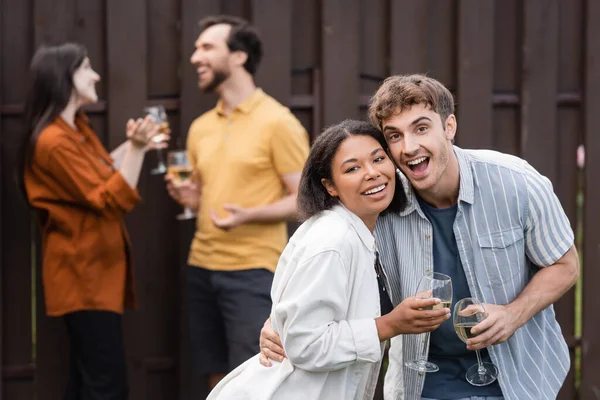 Alegre interracial pareja celebración vasos con vino cerca borrosa amigos en borrosa fondo - foto de stock