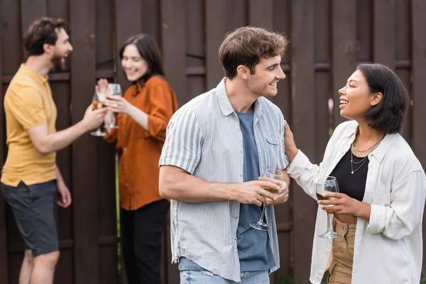 Cheerful interracial couple holding glasses with wine while talking near blurred friends on blurred background — Stock Photo